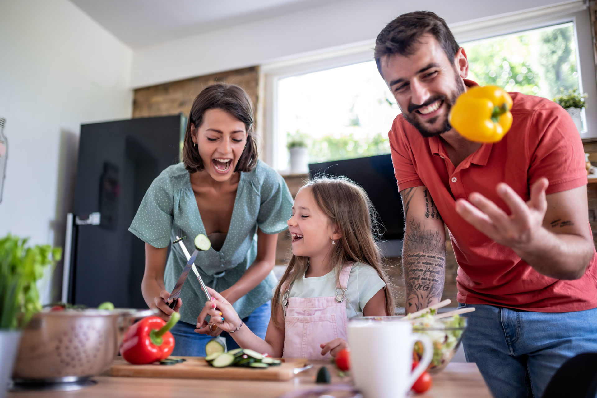 Familie hat Spaß beim Kochen in der Küche. Vater wirft gelben Paprika in die Luft. Mutter und Tochter spießen mit ihren Messern frisch geschnittenes Gemüse auf.