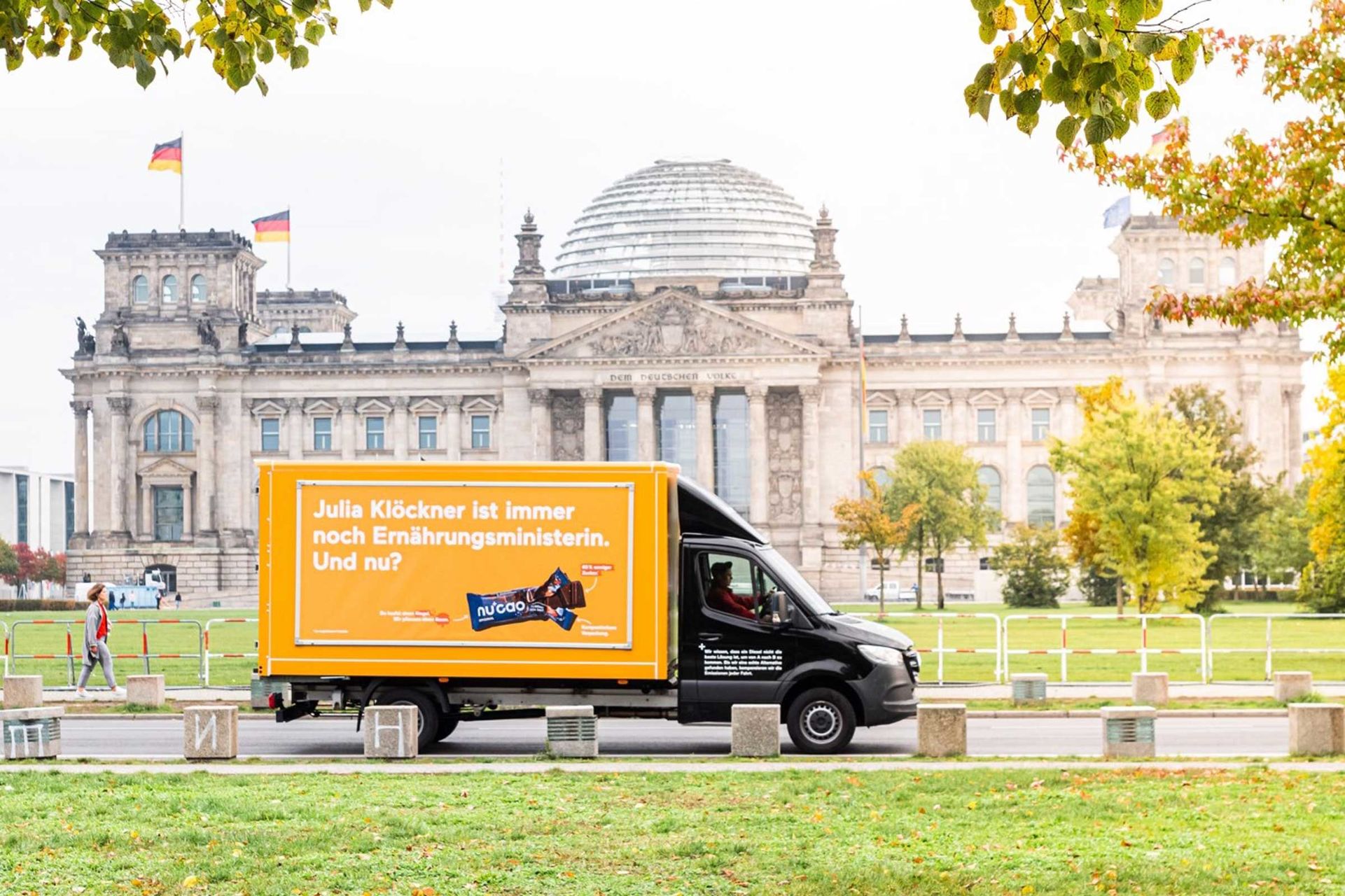 Schwarzer Kleintransporter mit Werbeplakat in orange auf der Ladefläche steht auf der Straße vor dem Reichstagsgebäude. Auf dem Werbeplakat steht „Julia Klöckner ist immer noch Ernährungsministerin. Und nu?