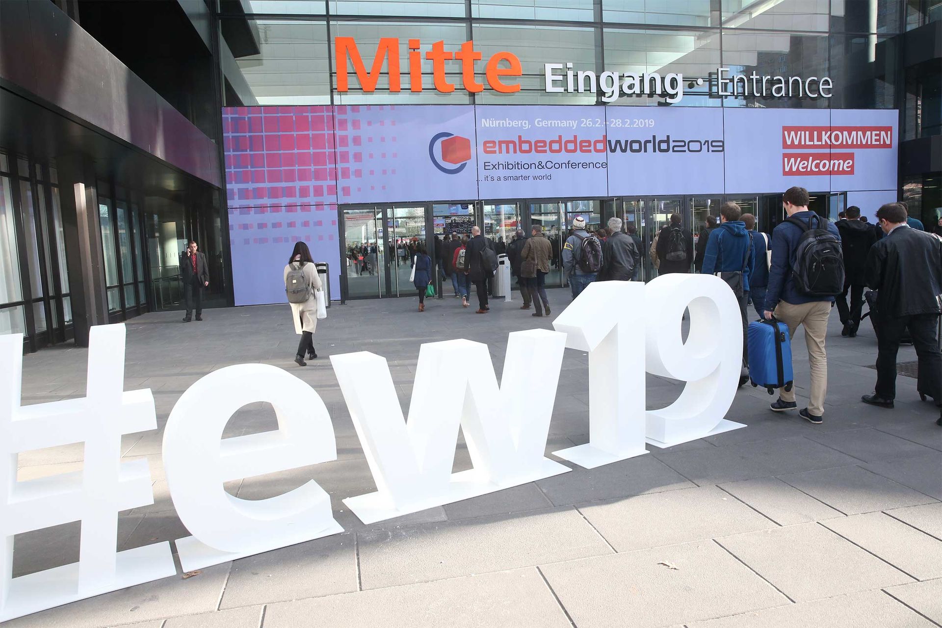 Das Foto zeigt sehr viele Menschen, die zur embedded world kommen und Richtung Eingangsbereich Mitte gehen. Im Vordergrund ist zudem der Hashtag ew19 zu sehen.