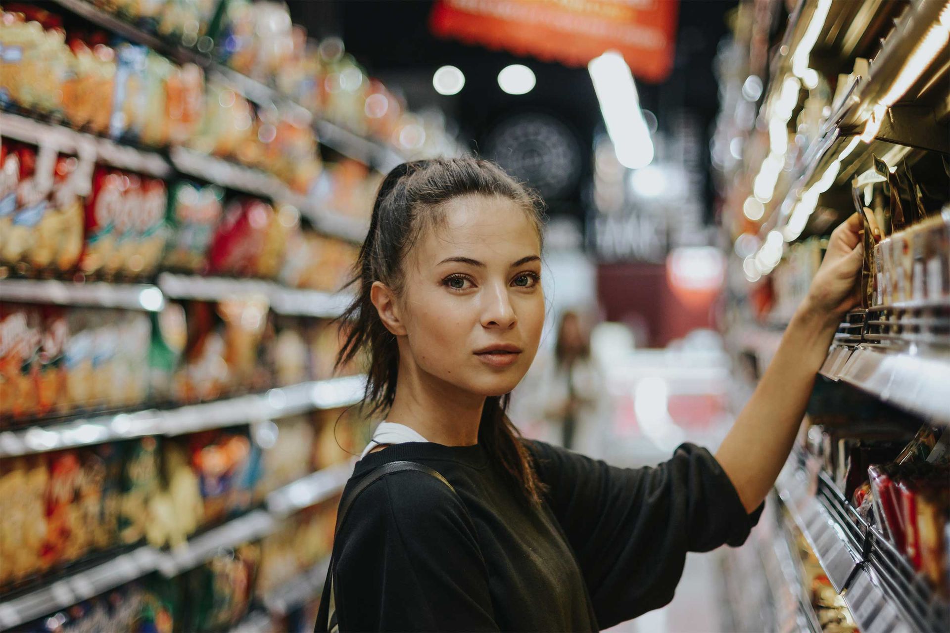Ein junge Frau in einer Supermarkt-Regalreihe greift nach einem verpackten Produkt und blickt in die Kamera.