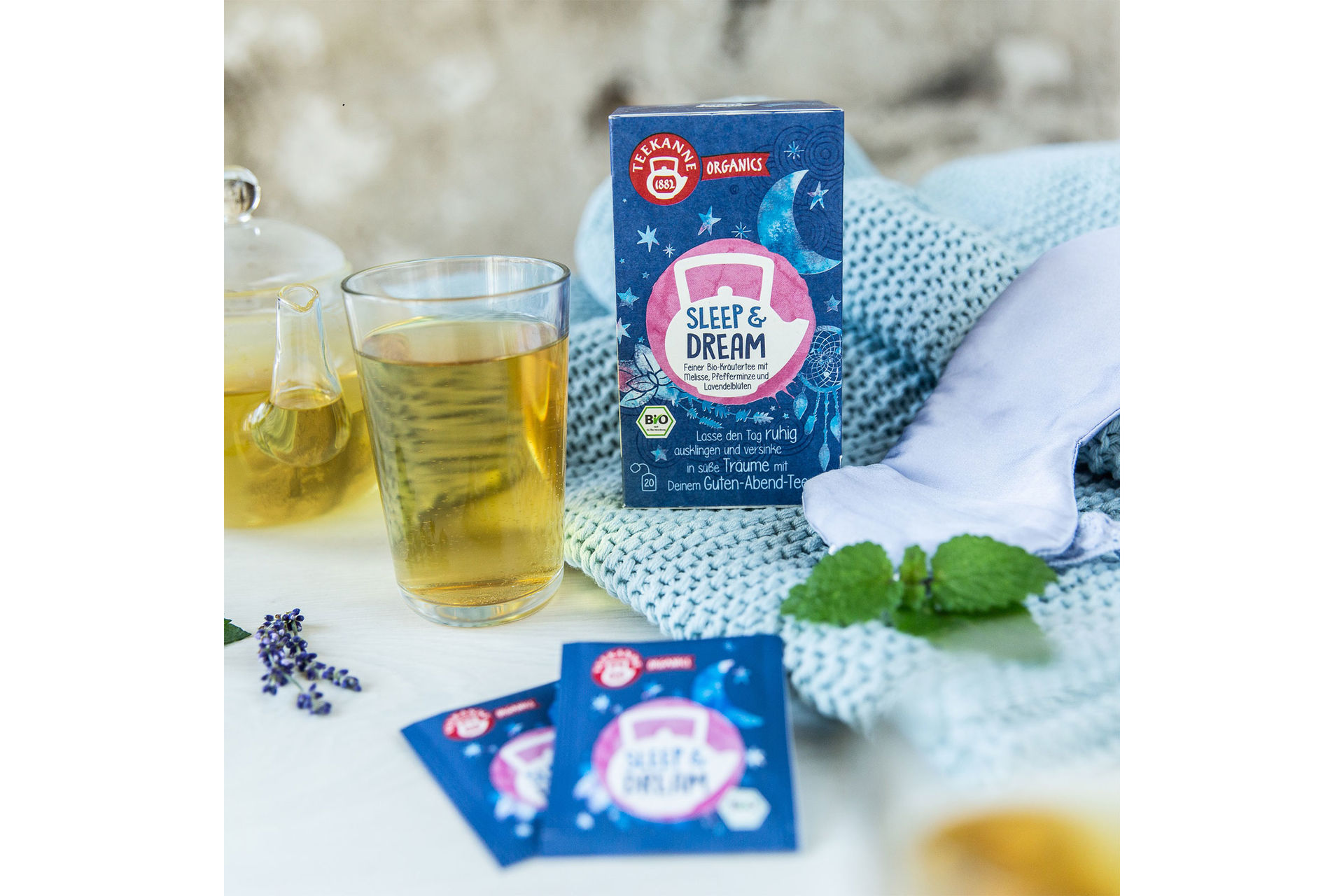 Stimmungsbild mit Kräutertee in einem Glas und einer Teekanne. Daneben liegen Teebeutel und eine Schachtel mit Teekanne Organics Tee.