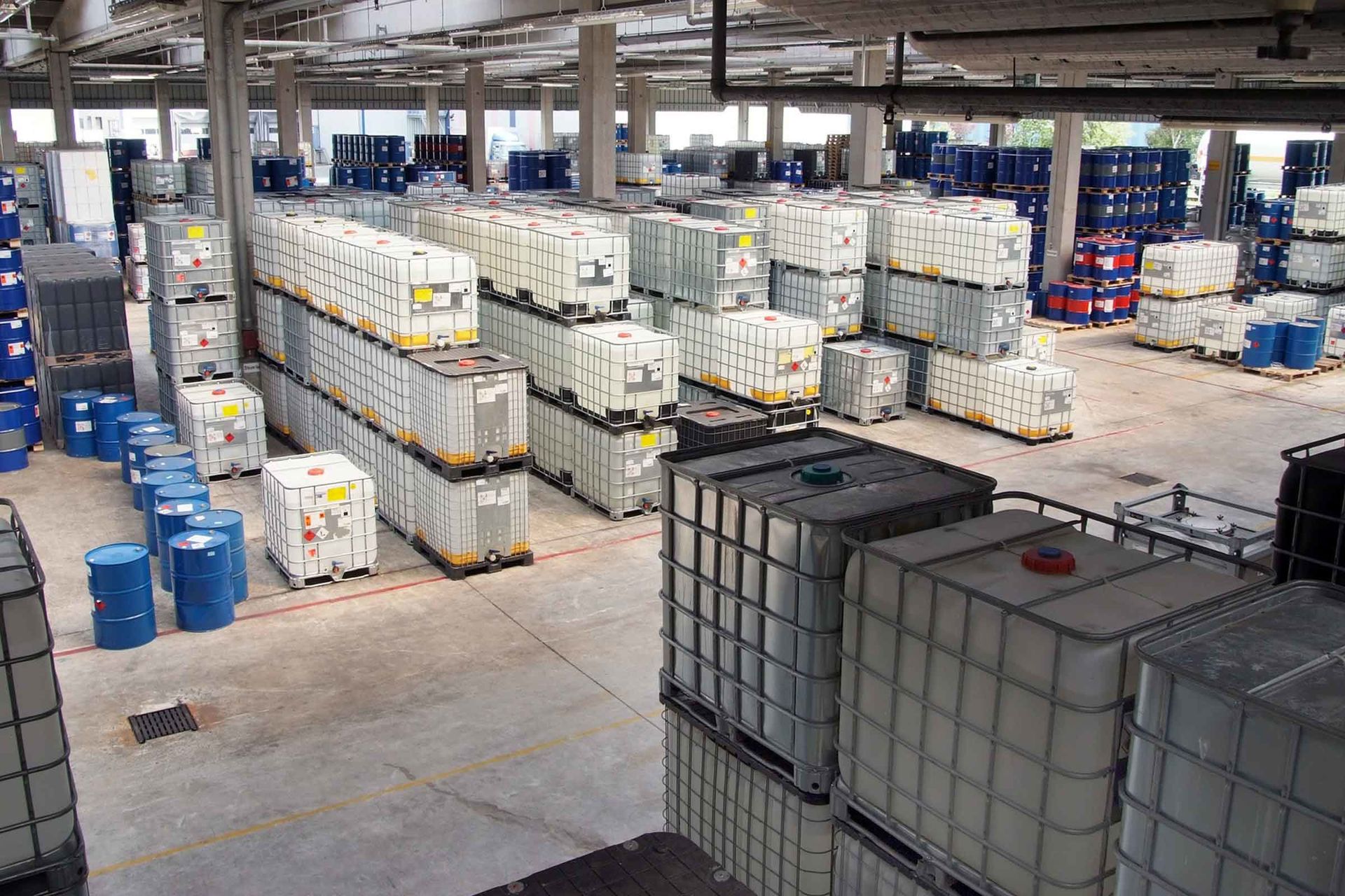 Ein Chemielager mit gestapelten IBC-Containern aus Kunststoff und Metall und blaue Metallfässer mit Gefahrenhinweisen