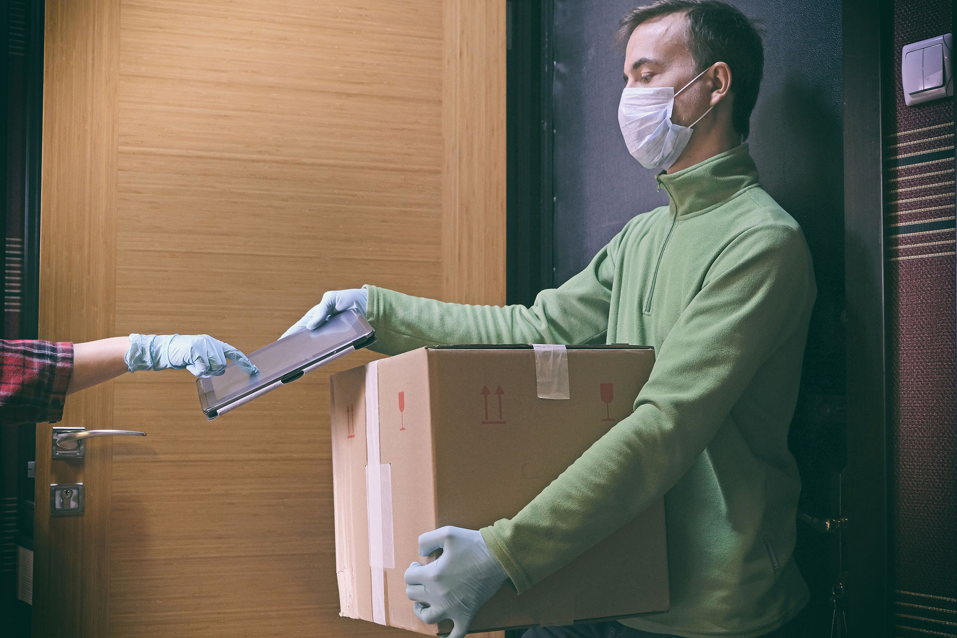 Ein Paketlieferant mit Schutzmaske und Latexhandschuhen hält vor einer Haustüre einen Karton in der Hand. Die Kundin quittiert den Empfang auf einem Tablet - von ihr ist nur die Hand im Latexhandschuh zu sehen.