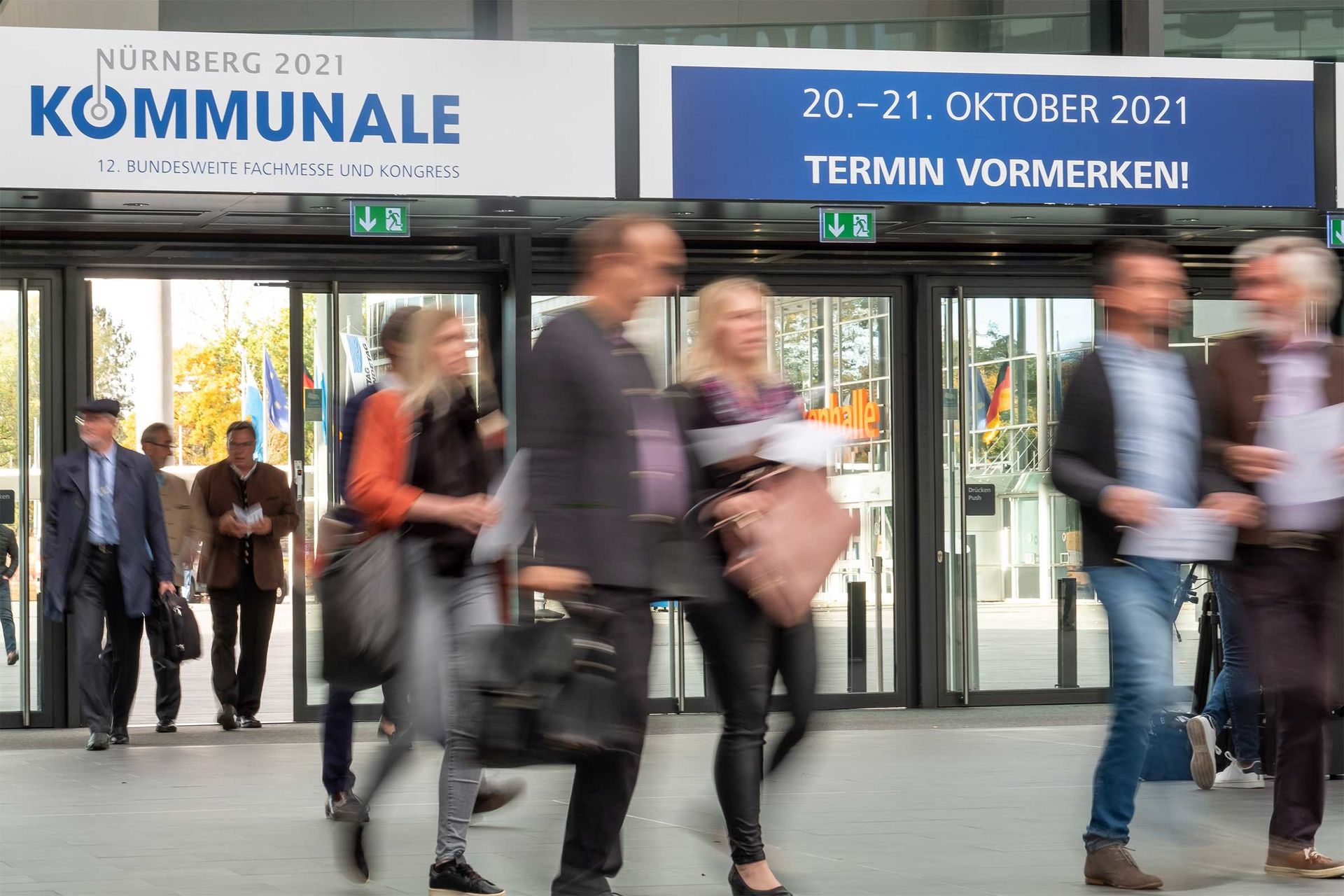 Das Bild zeigt Menschen, die in die Messe laufen und das Datum der nächsten KOMMUNALE: vom 20.- 21. Oktober 2021