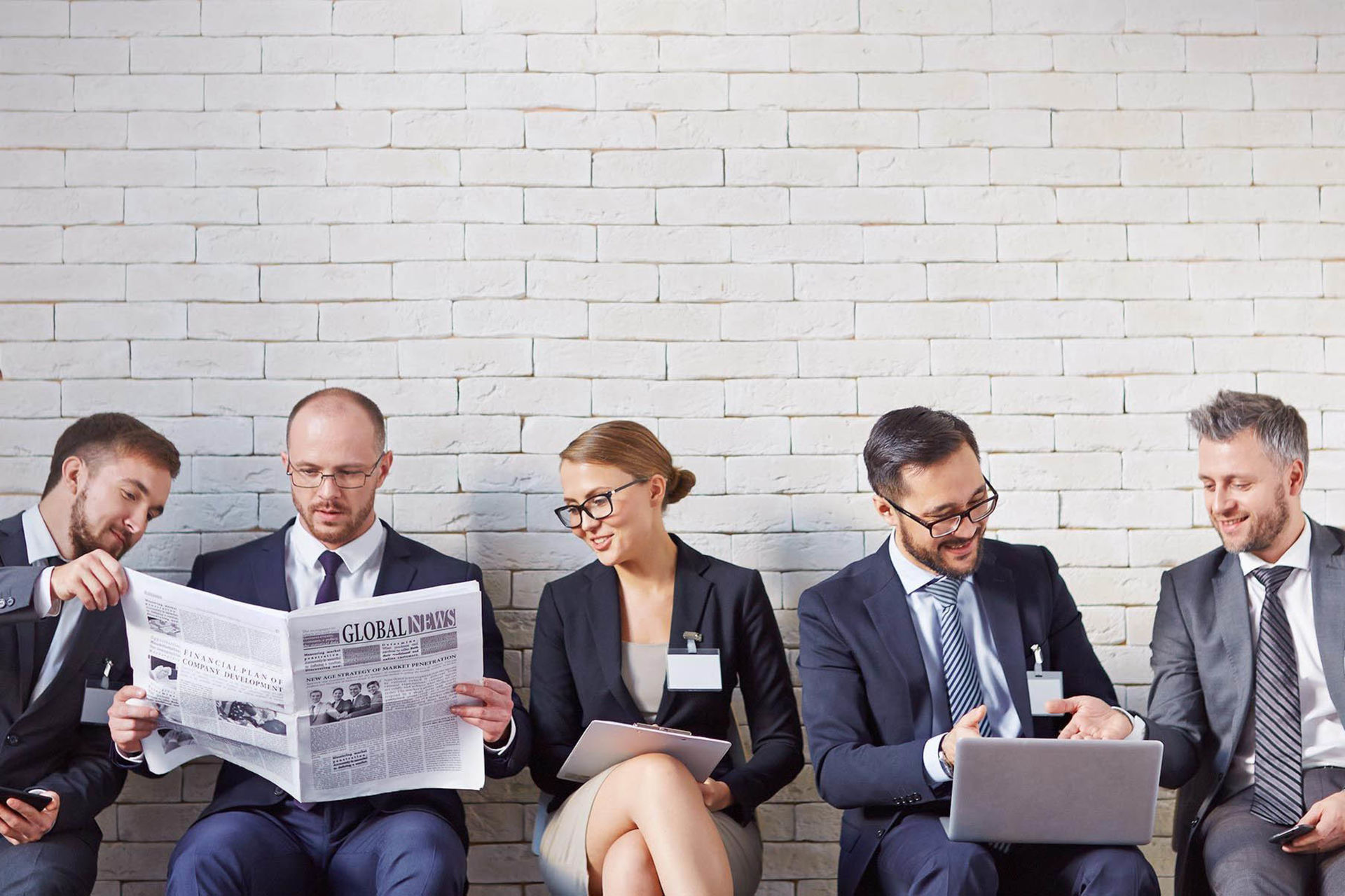 5 Personen sitzen auf einer Bank, lesen Zeitung und schauen ins Laptop