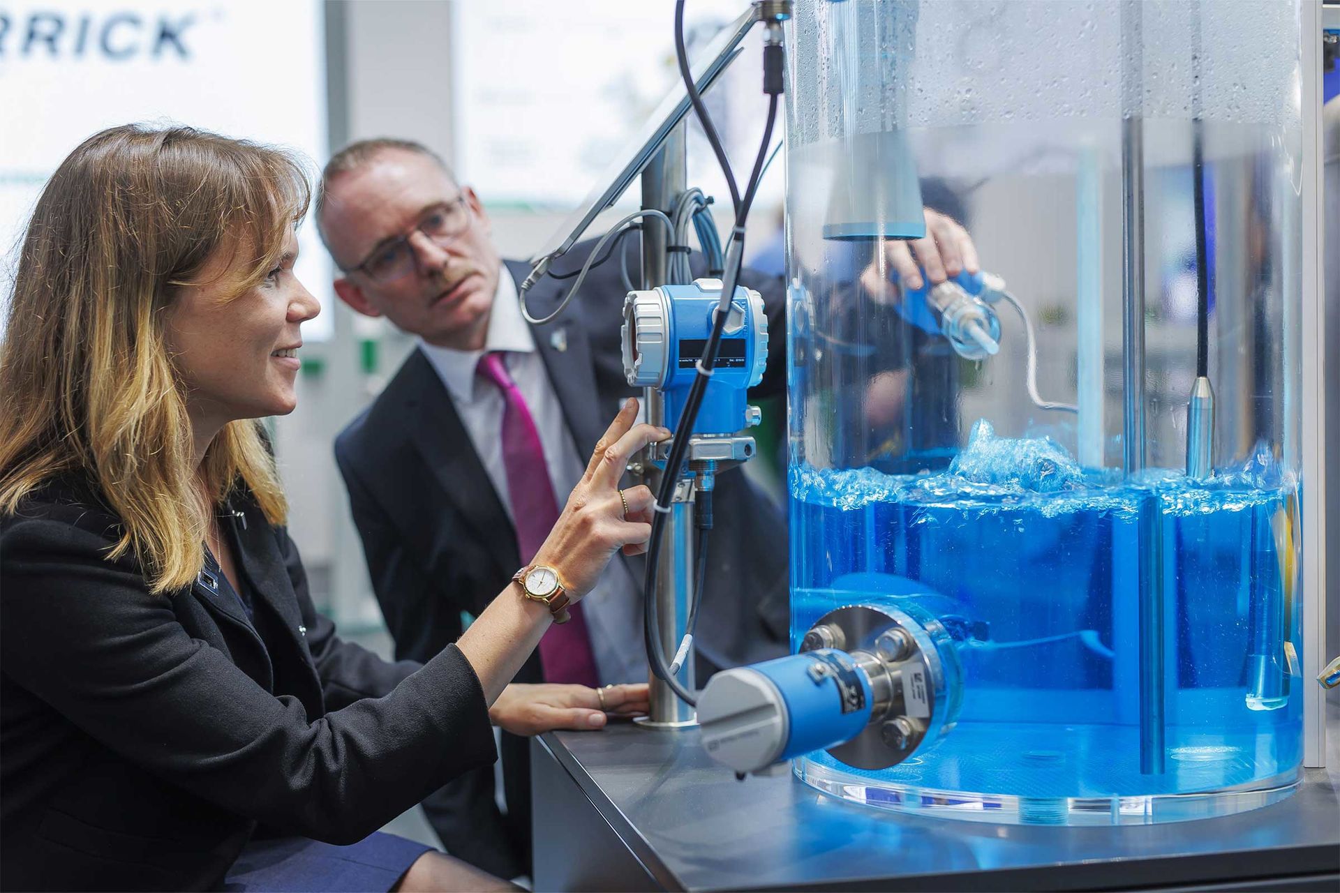 Eine Frau zeigt einem Mann auf der POWTECH eine Maschine, die Flüssigkeit verarbeitet.