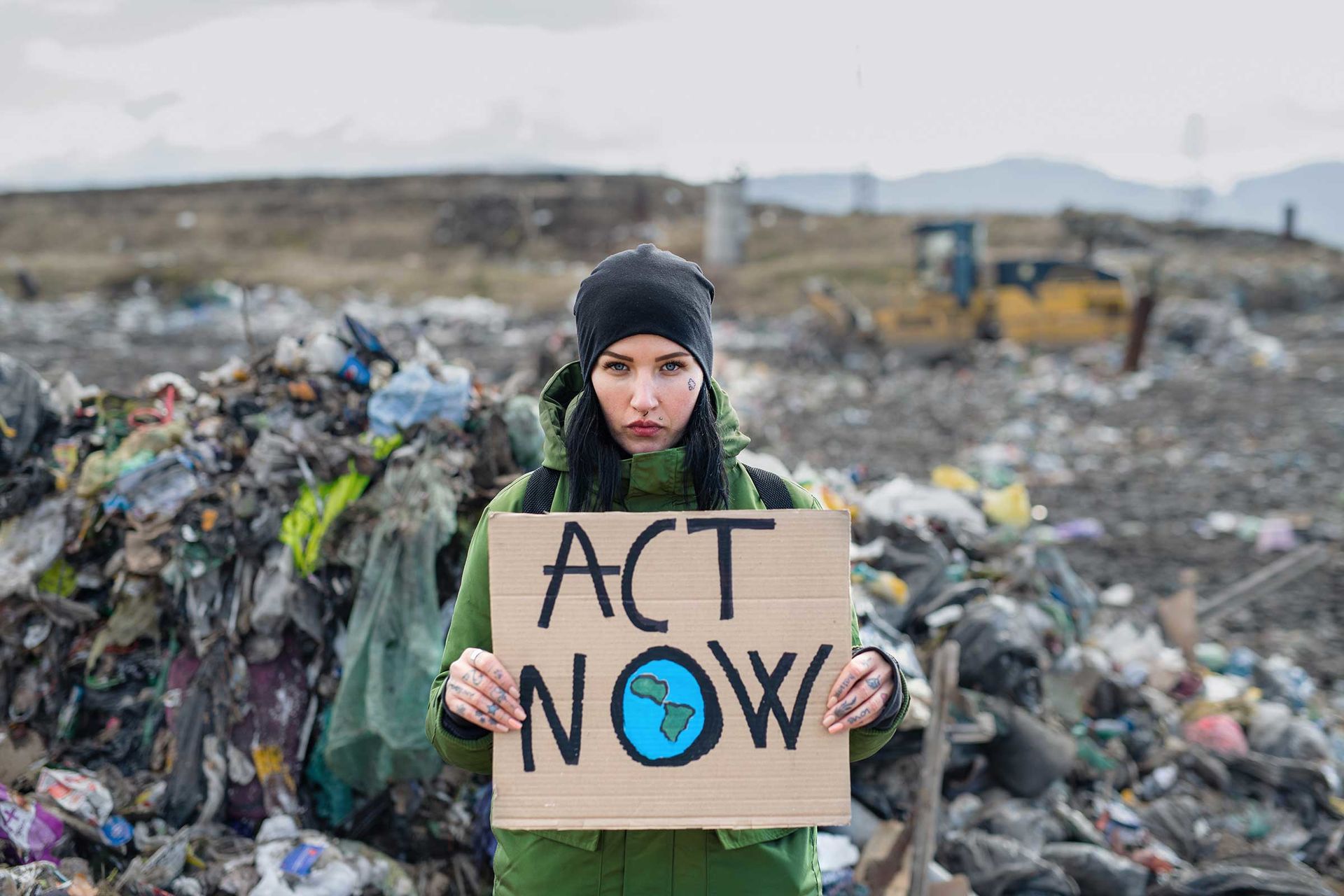 Junge Frau steht mit ernstem Blick vor einer Müllhalde, in der Hand ein Pappschild mit der Aufschrift „Act now“