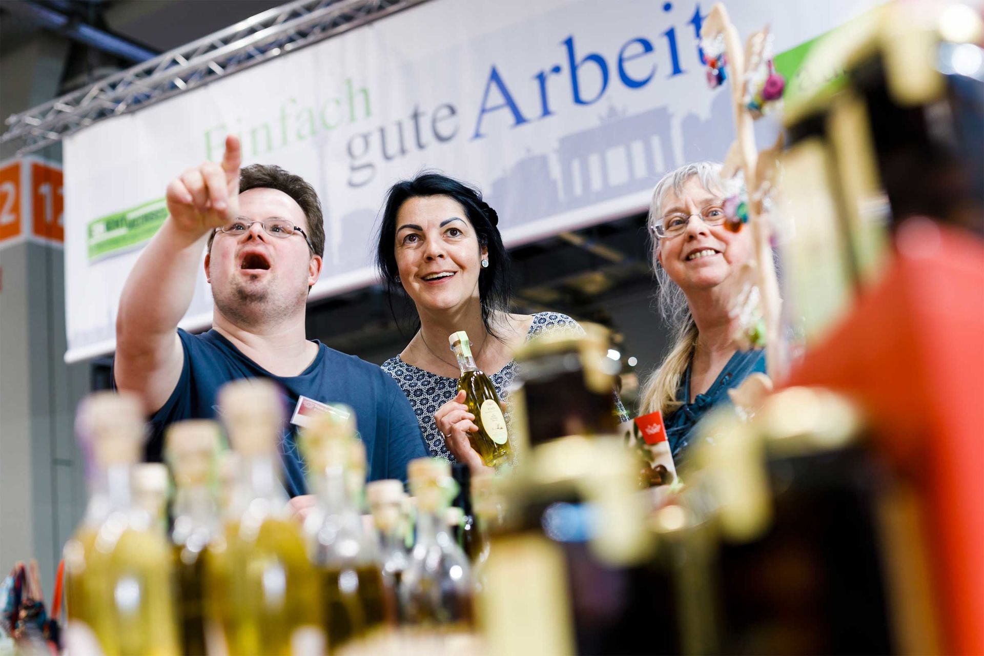 Das Bild zeigt einen Mann und zwei Frauen voller Freude auf der Werkstätten:Messe. Der Mann zeigt mit dem Finger auf etwas in der Ferne und die Frauen schauen begeistert hin.
